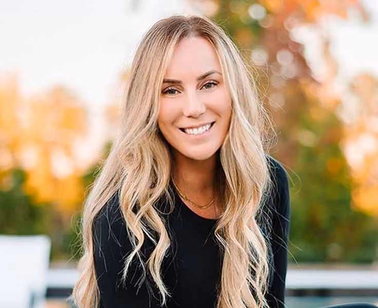 Smiling woman with long wavy blonde hair wearing a black top, outdoors with blurred greenery and autumn colors in the background.