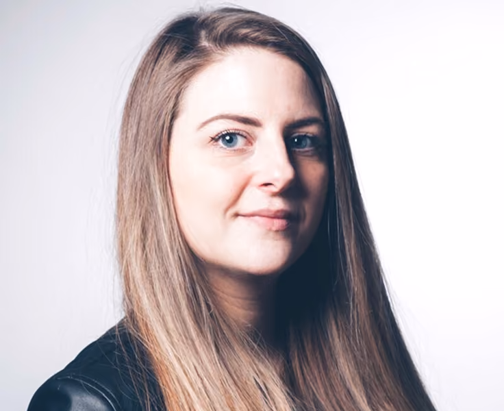 Portrait of a woman with long straight light brown hair and blue eyes, wearing a black top, against a white background.