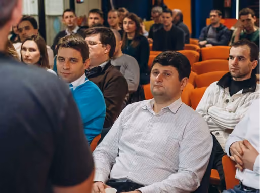 Audience of diverse adults sitting in orange chairs attentively listening to a speaker who is partially visible from behind in the foreground.