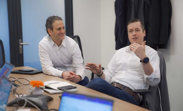 Two men in white shirts sitting and talking in an office meeting room with laptops and notebooks on the table.