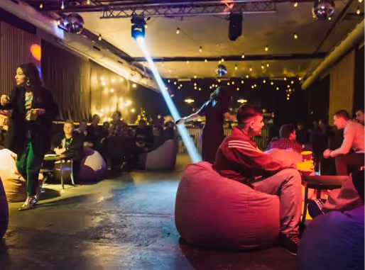 A dimly lit social lounge with people sitting on bean bags and chairs, warm string lights on the walls, and a bright spotlight illuminating the ceiling.