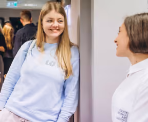 Two women smiling and talking indoors; one in a light blue sweatshirt leaning against a wall, the other facing her in a white shirt.