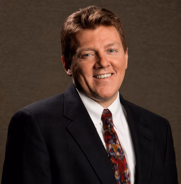 Smiling middle-aged man with light brown hair wearing a black suit, white shirt, and multicolored tie standing against a plain dark brown background.