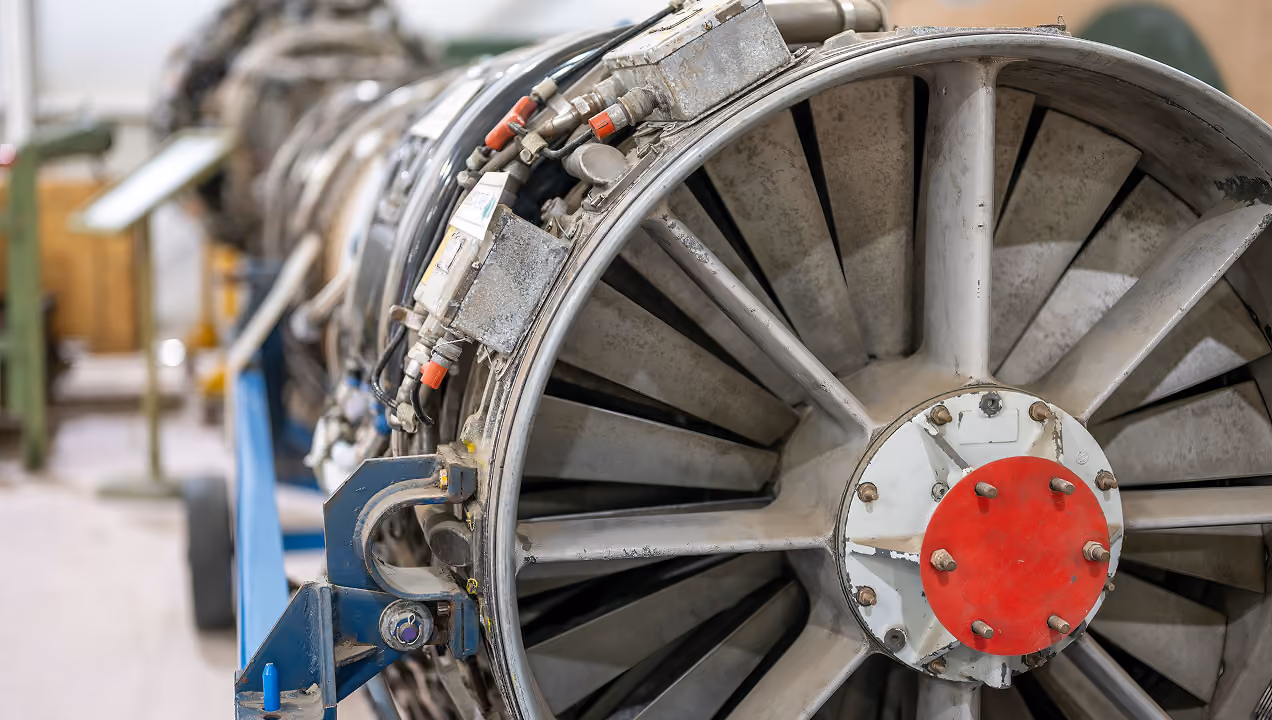 Close-up view of a large industrial turbine or jet engine component with visible fan blades and connected mechanical parts in a workshop setting.