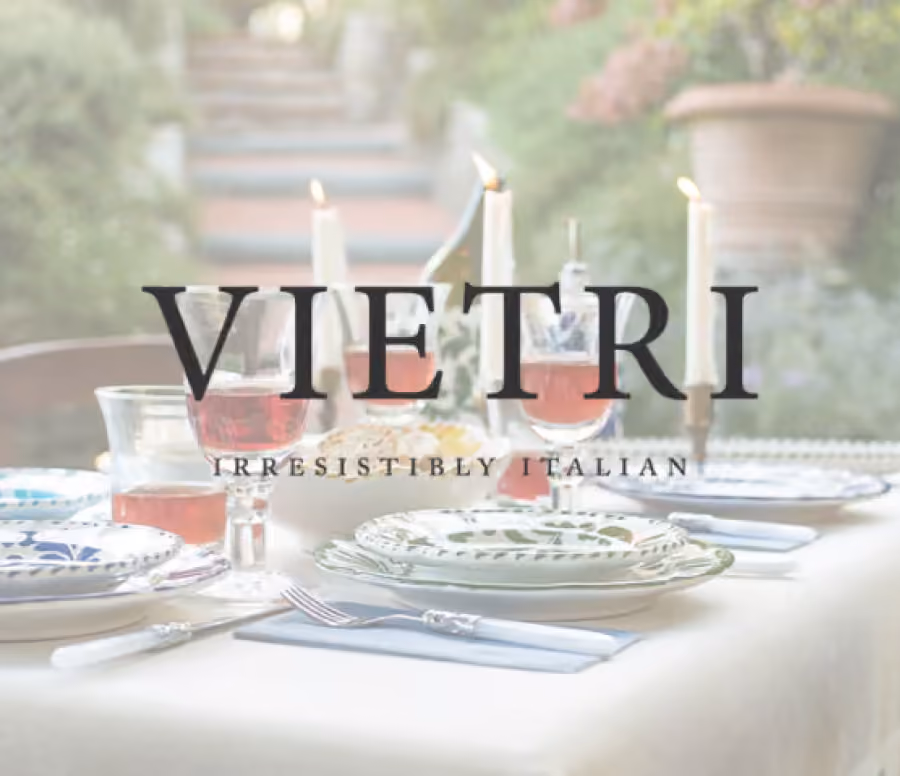 Outdoor dining table set with patterned plates, silverware, four glasses of rosé wine, lit white candles, and a white tablecloth, with a garden background.