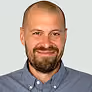 Smiling bald man with a beard wearing a blue button-up shirt against a white background.