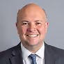 Smiling bald man in a suit and tie against a plain gray background.