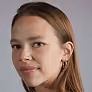 Portrait of a woman with light skin and brown hair, wearing a black top and looking slightly to the right with a neutral expression, against a plain gray background.