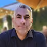 Middle-aged man with short salt-and-pepper hair and a dark collared shirt standing outdoors in warm lighting with blurred trees and string lights in the background.