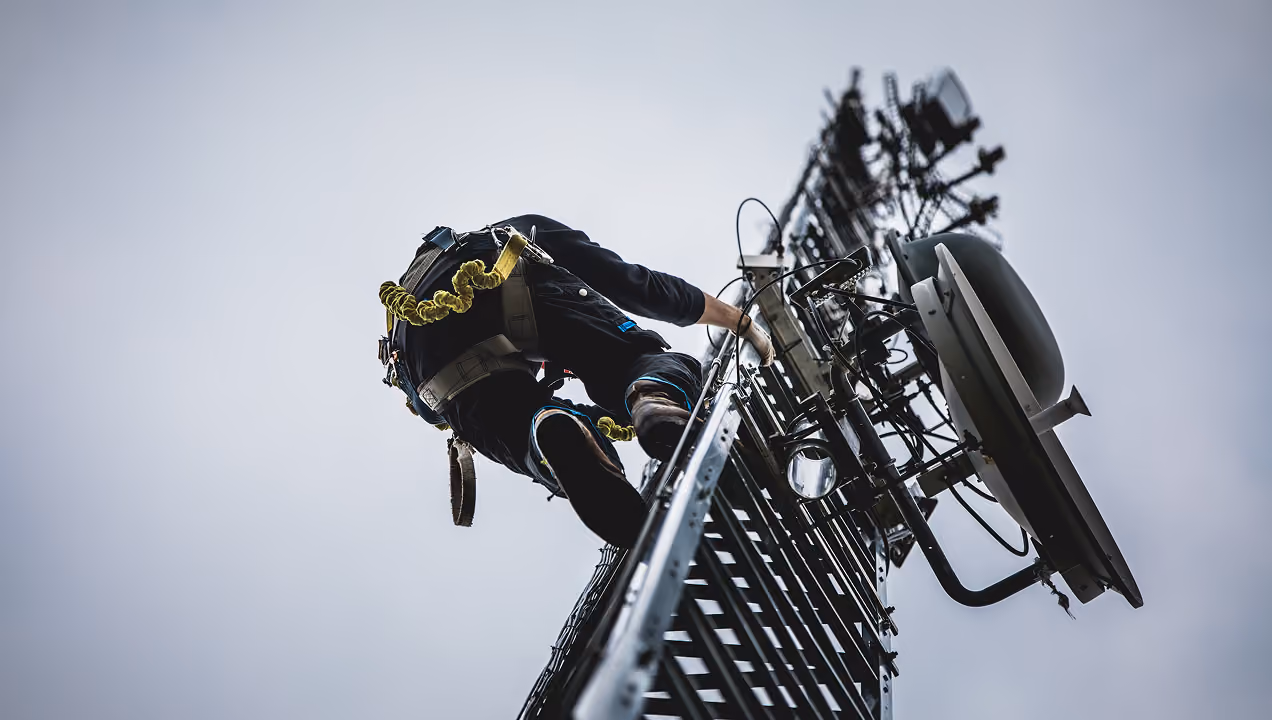 Technician climbing a telecommunications tower wearing safety gear and harness, with antennas and cables attached to the structure against a clear sky background.