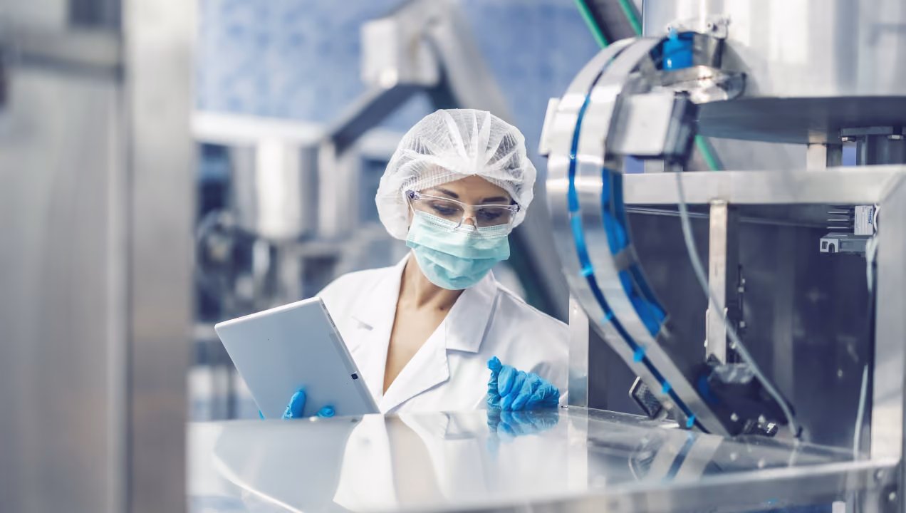 A medical professional wearing protective gear including a hairnet, face mask, safety glasses, and gloves, using a tablet while inspecting machinery in a sterile industrial or laboratory setting.