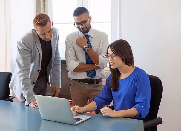 Three business professionals, one woman sitting and two men standing, collaborating and looking at a laptop screen in an office setting with a window in the background.