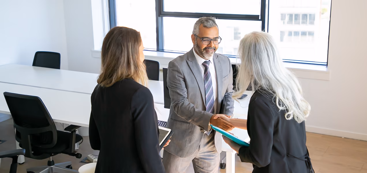Three business professionals in an office with large windows, two women and one man, shaking hands and smiling during a meeting, with empty desks and chairs in the background.