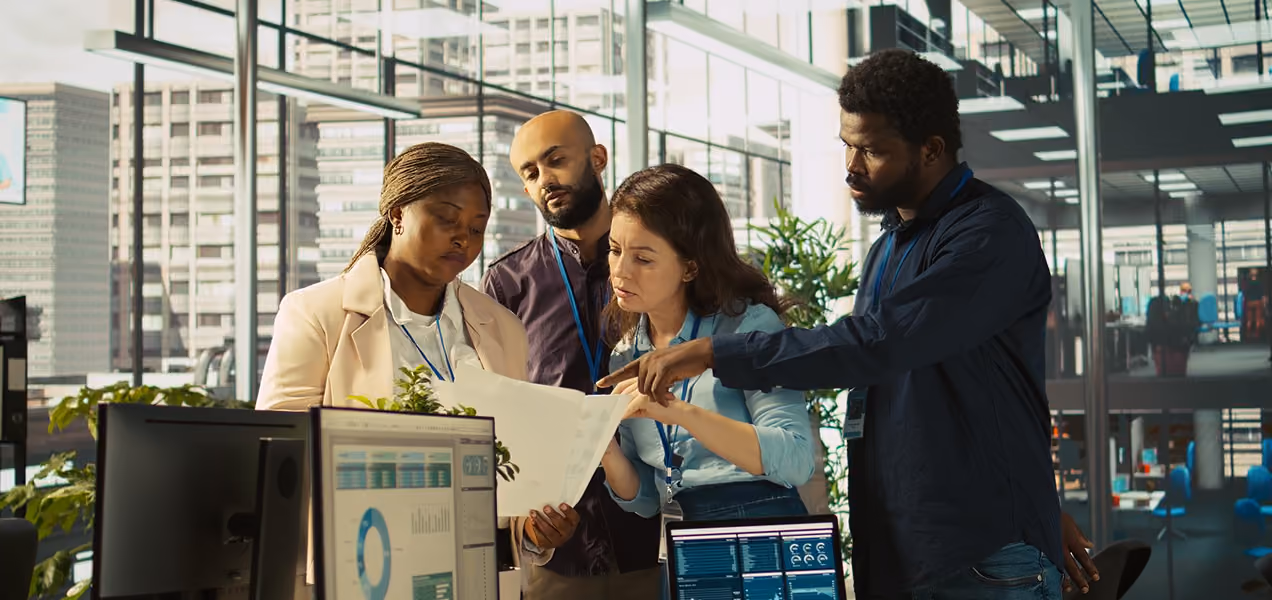 Four diverse coworkers gathered in an office with glass walls, examining documents together with computer screens displaying data charts in the foreground.