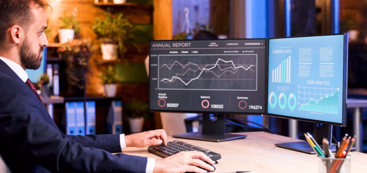 Man in business suit working at a desk with dual monitors showing business data visualizations including a line graph labeled Annual Report, bar charts, and pie charts.