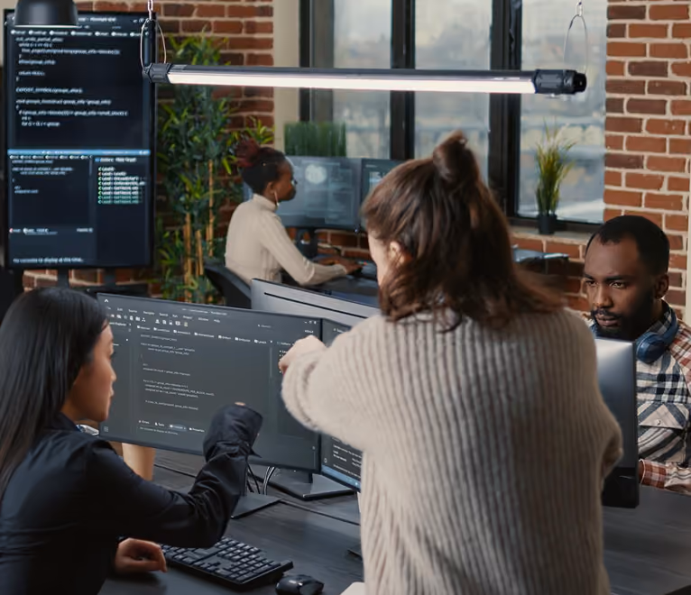 Four people working in a modern office with exposed brick walls and large windows; two women in the foreground collaborate over computer code displayed on dual monitors, while a man and another woman work independently at their desks in the background.