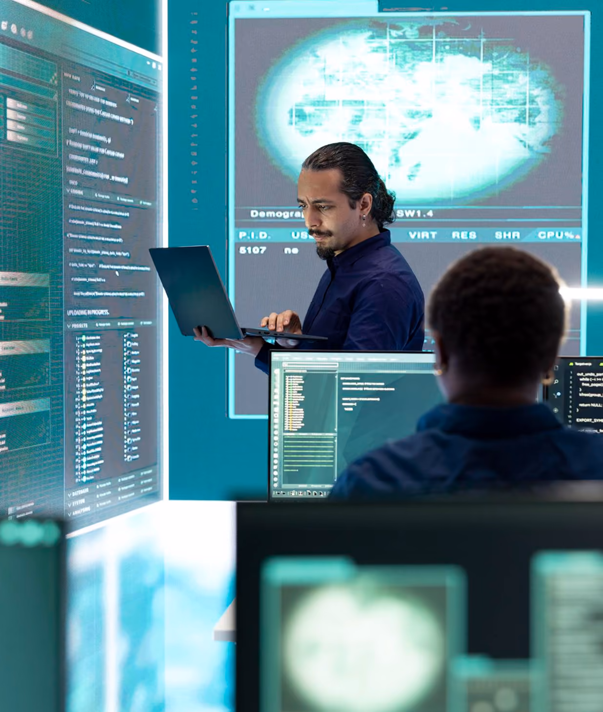 Two men in a technology workspace with multiple computer screens showing code and a world map; one man stands holding a laptop, the other sits facing monitors.
