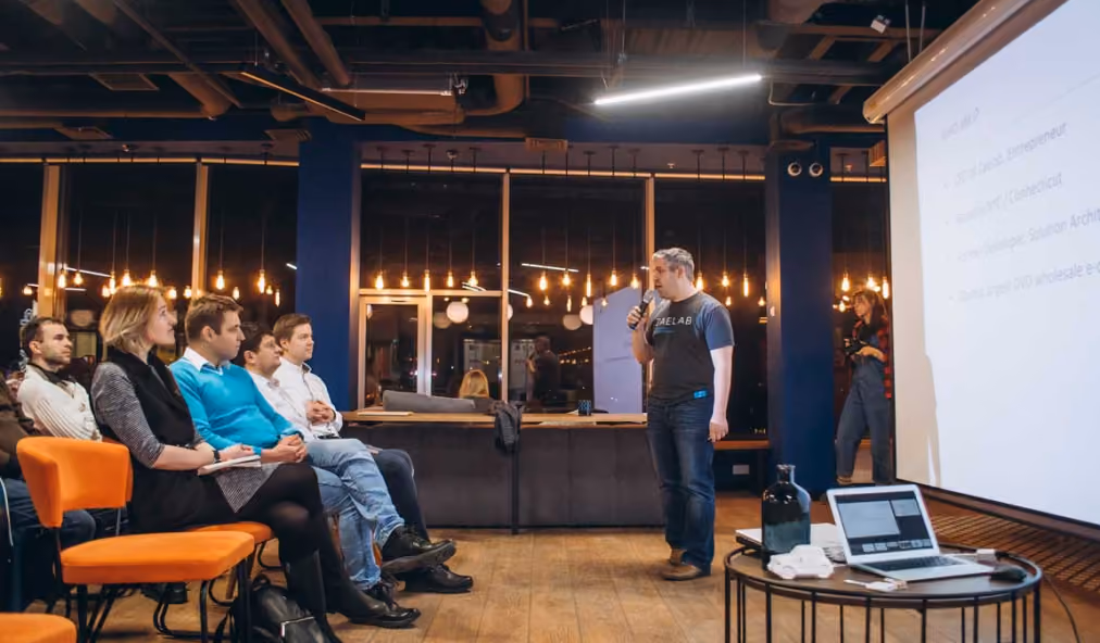 A man in a black T-shirt and jeans holds a microphone and speaks in front of a seated audience in a modern room with wooden floors, warm hanging lights, and a large projected screen. A laptop and a black bottle are on a small round table near the screen.