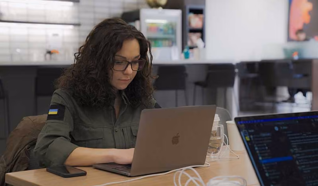 Woman with curly dark hair and glasses, wearing a green jacket with a Ukrainian flag patch, working on a laptop at a wooden table in a modern office setting.