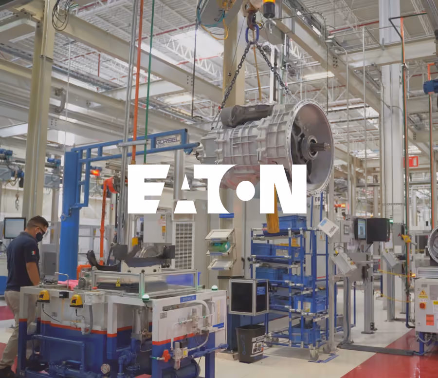 Industrial factory floor with a large transmission unit suspended by a chain hoist, machinery and a worker wearing a face mask on the left side, with Eaton logo overlay.