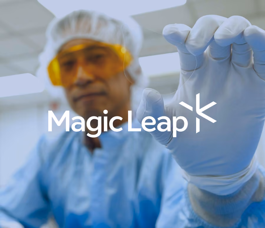 Scientist in a cleanroom suit and yellow safety glasses holding a thin, transparent flexible electronic component with gloved hands.