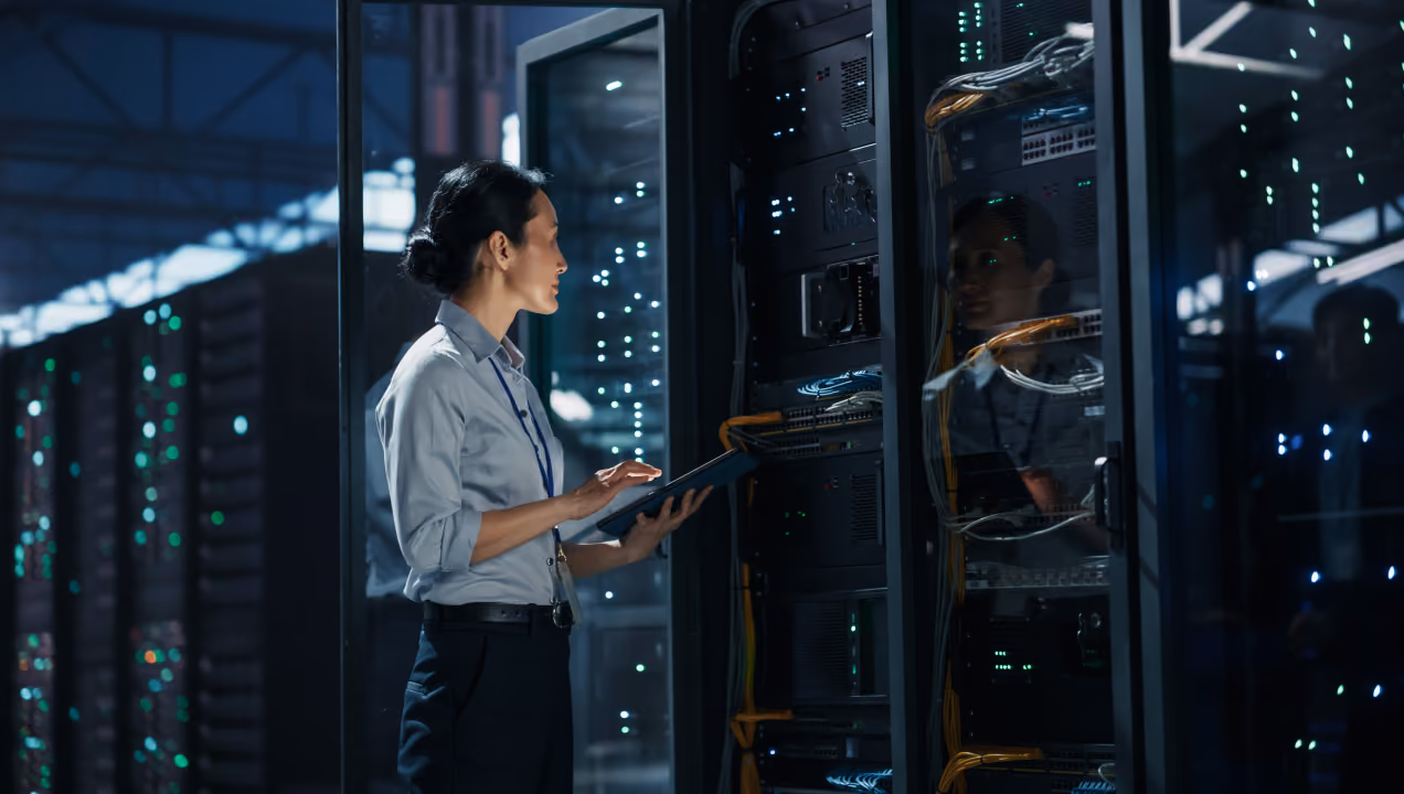 Woman in a light blue shirt holding a tablet and working in a data center with server racks and network cables.