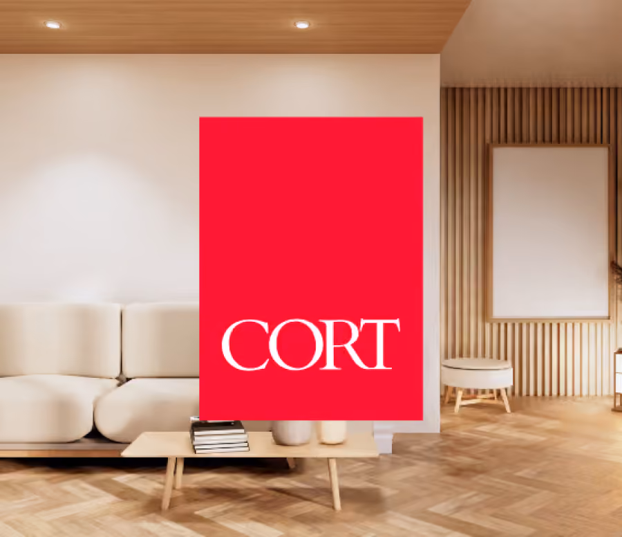 Modern living room with light wood flooring, beige sofa, wooden coffee table holding stacked books, and a red vertical rectangular sign with the word 'CORT' in white letters in the center.