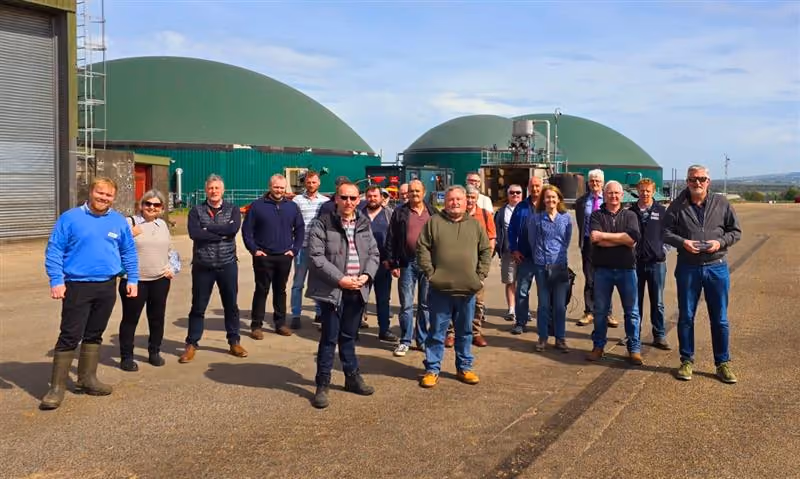 Group of people standing outdoors on concrete with large green industrial tanks behind them.