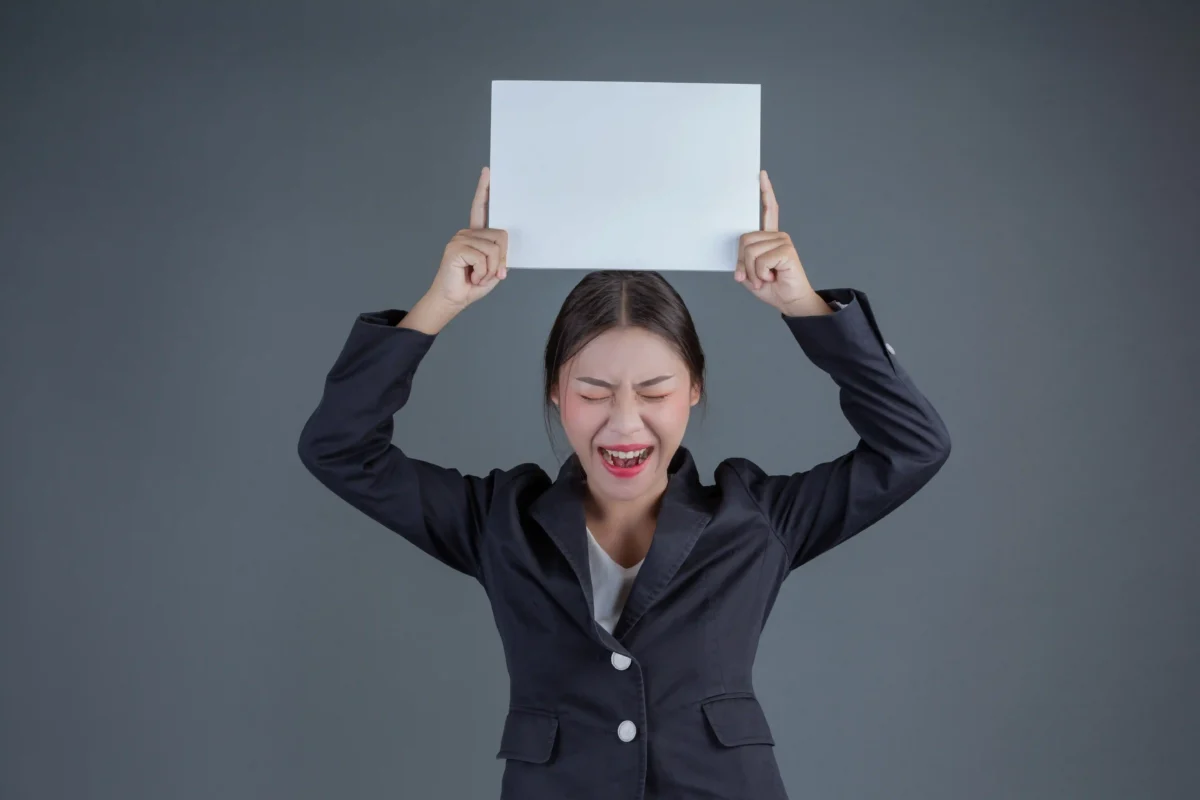 A woman in a business suit stands with her eyes closed and mouth open in a shout, holding a blank white board. The image conveys a feeling of unheard frustration and a desperate need to be acknowledged.