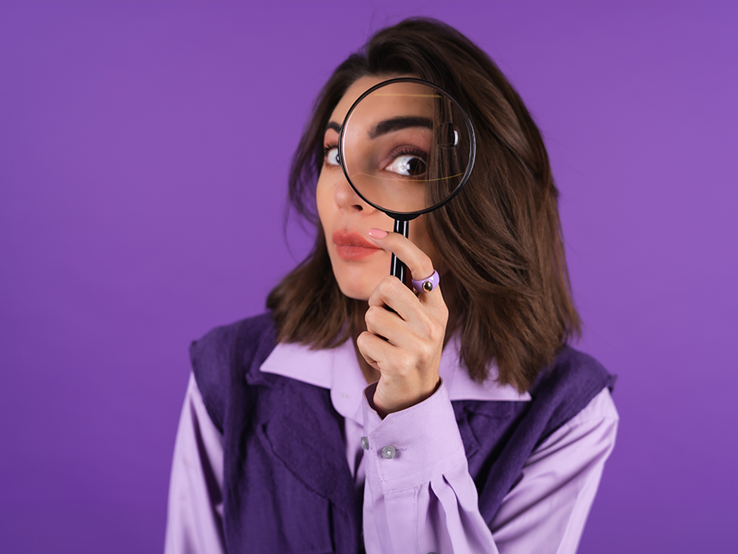 Woman in purple shirt holding a magnifying glass over one eye against a purple background.