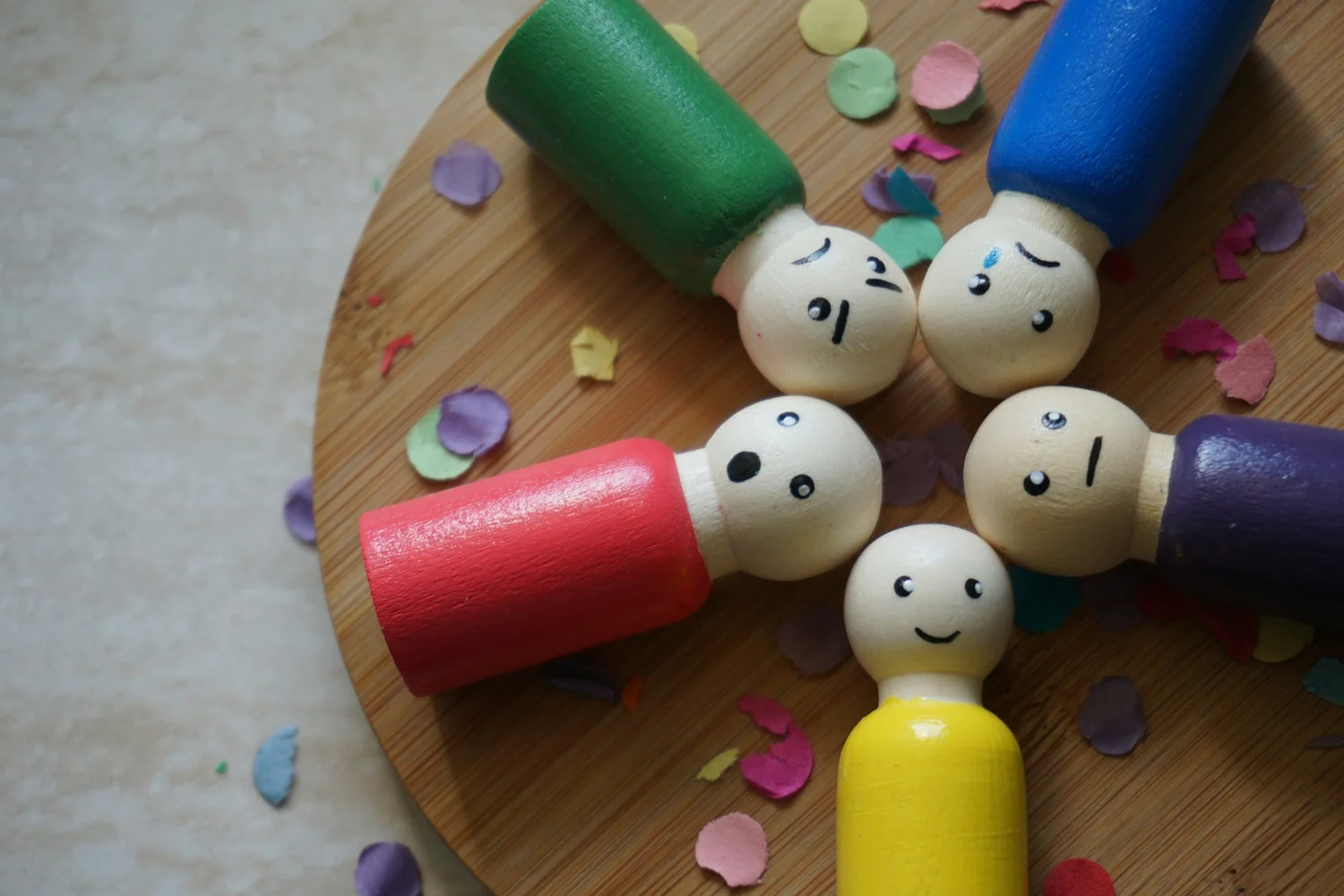 Five wooden peg dolls with different facial expressions arranged in a circle on a wooden board with colorful confetti