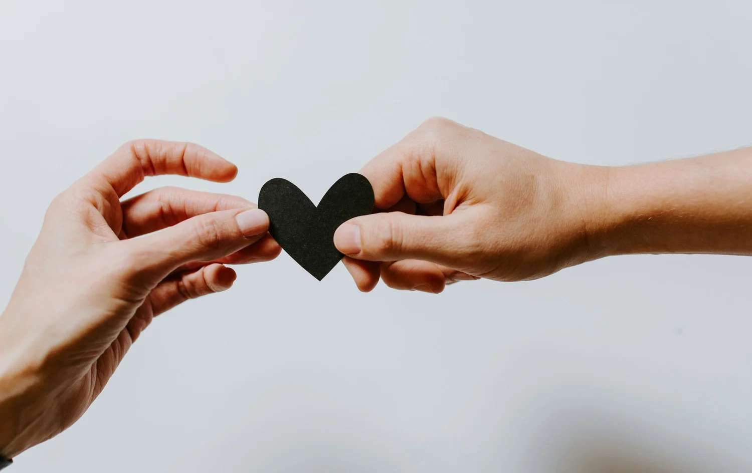 Two hands holding a small black paper heart between them against a white background