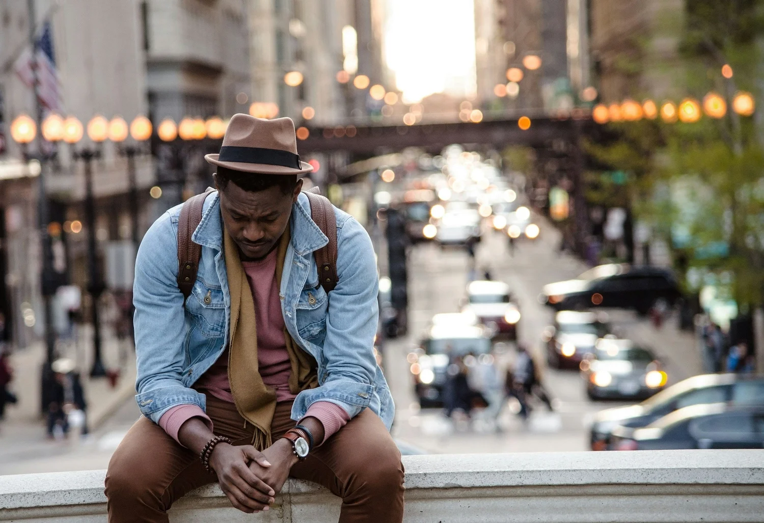 A person in a hat and denim jacket sits on a ledge looking down, hands clasped, above a busy city street
