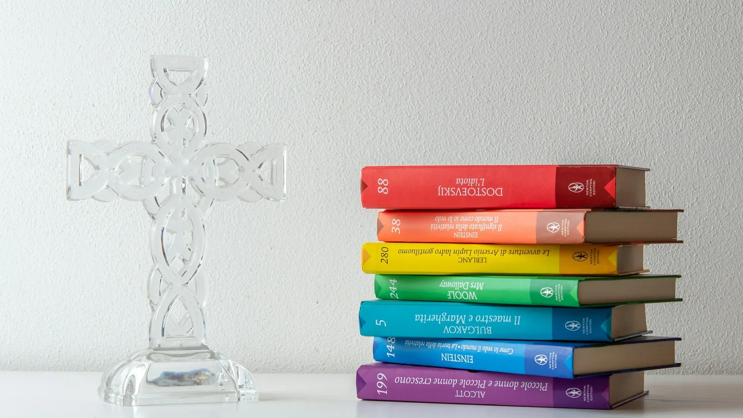 Clear glass cross with a knotwork design beside a stack of six books with rainbow-colored spines