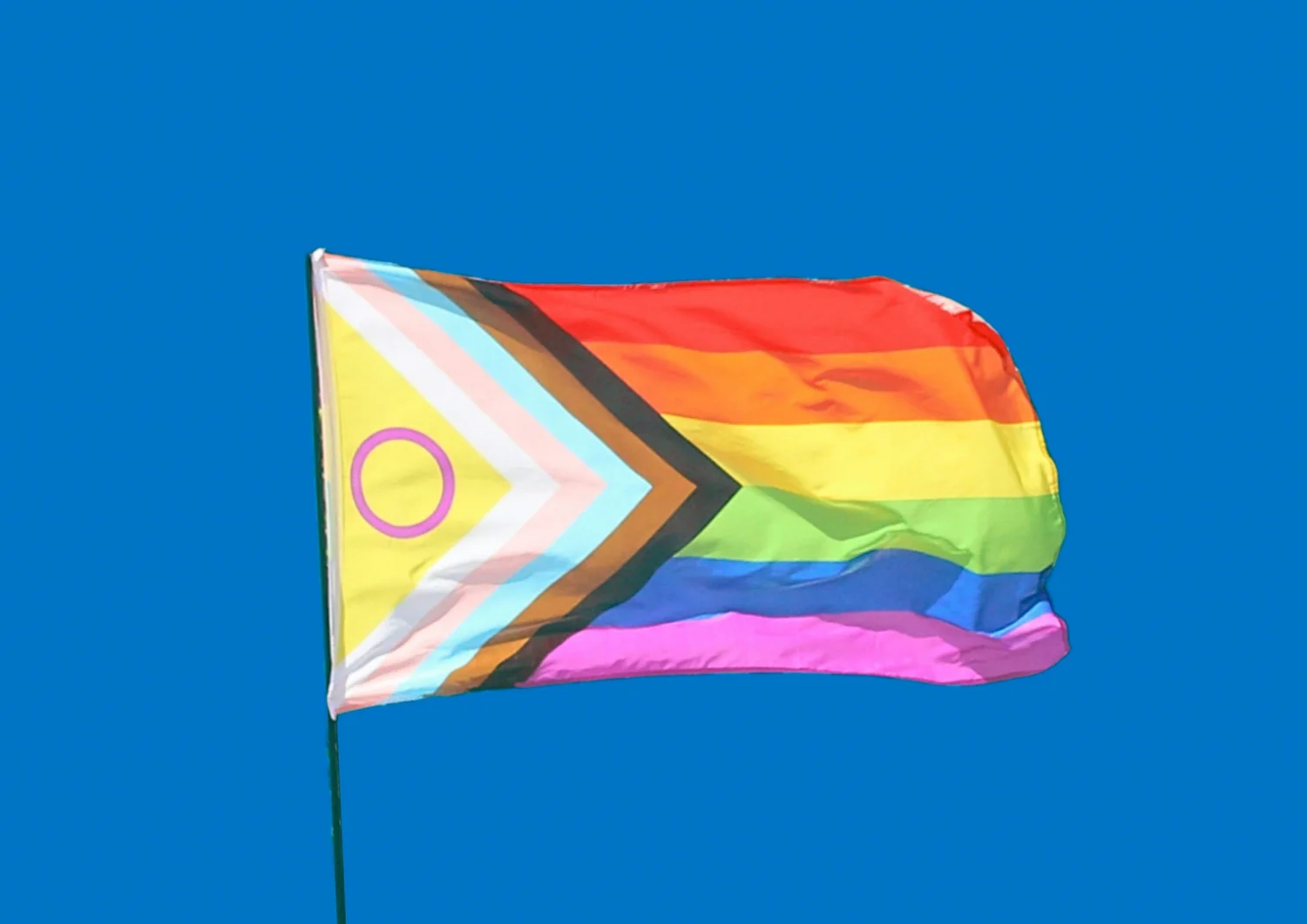Rainbow flag with a multicolored chevron and a purple ring on yellow, waving against a clear blue sky