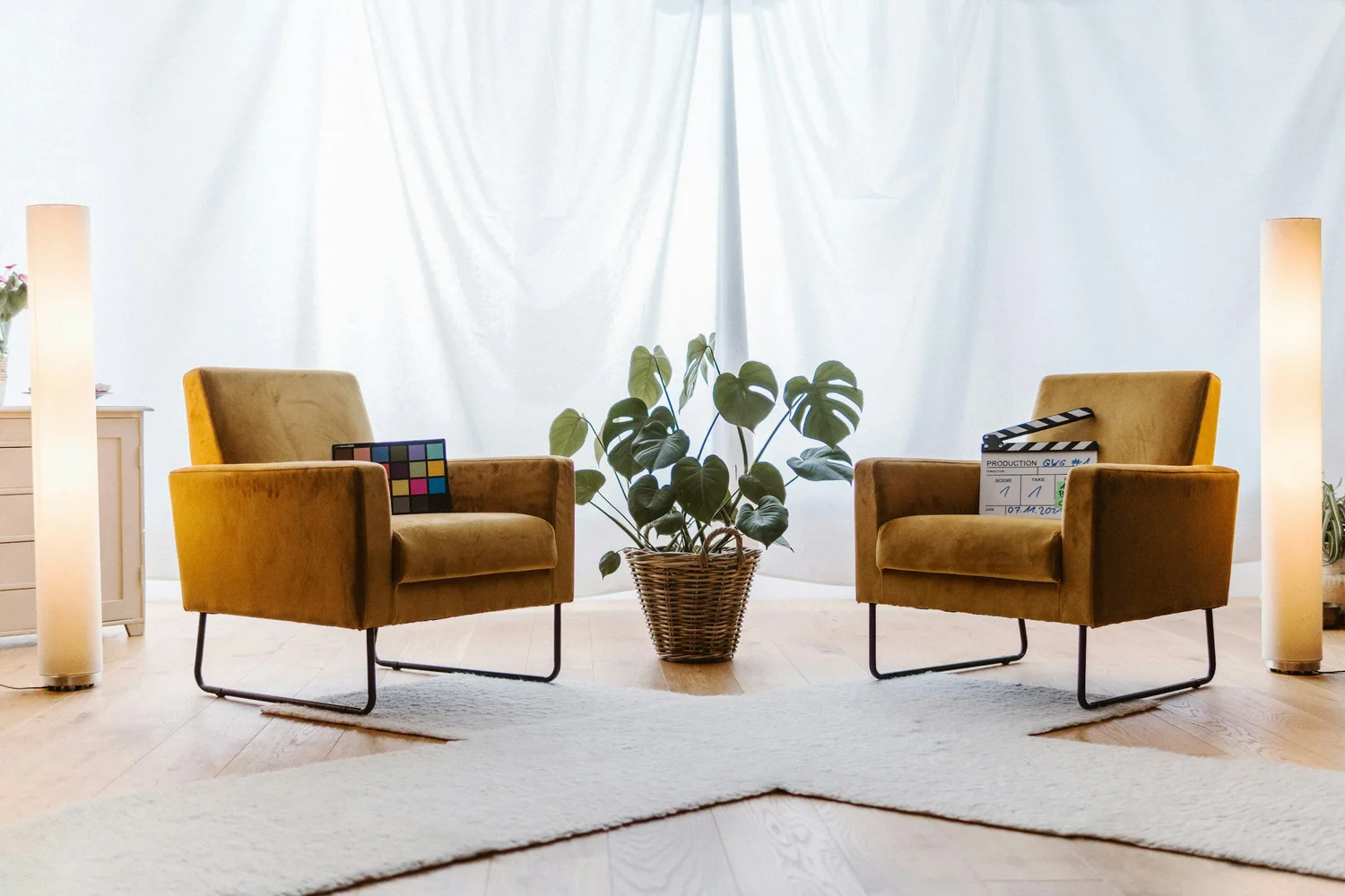 Two mustard armchairs facing each other around a potted plant in a basket, with white curtains and floor lamps