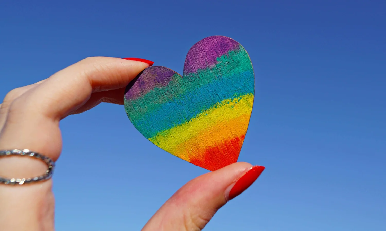 Hand with red nails holding up a rainbow-striped wooden heart against a clear blue sky