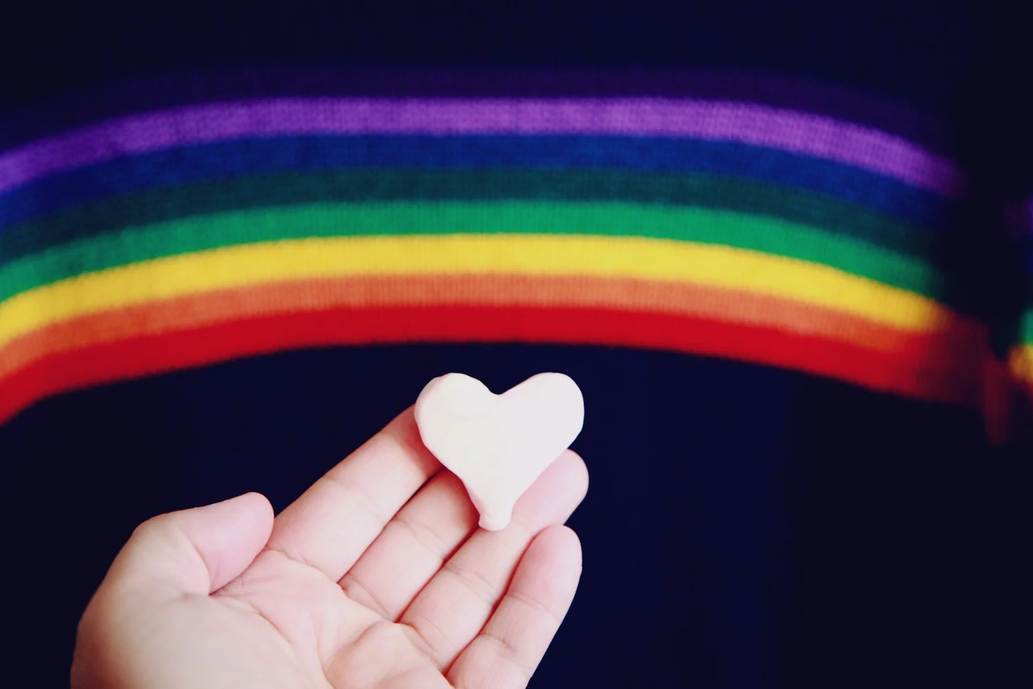 Open hand holding a small white heart cutout in front of a dark background with a rainbow arc