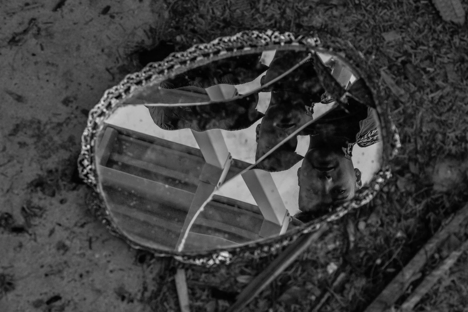 Cracked oval mirror in an ornate frame lying on the ground, in black and white