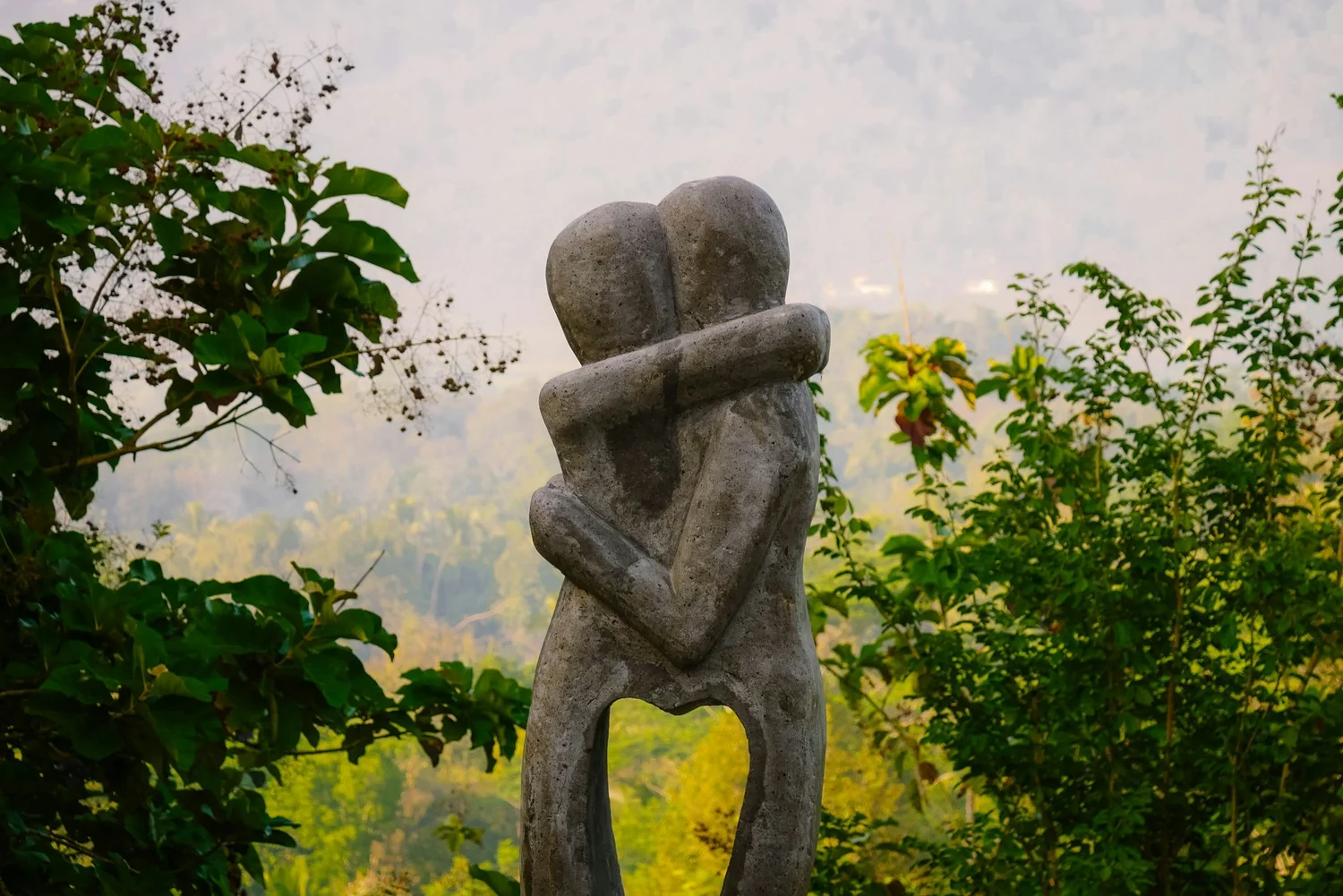 Carved sculpture of two figures in an embrace, set among green trees with misty hills behind