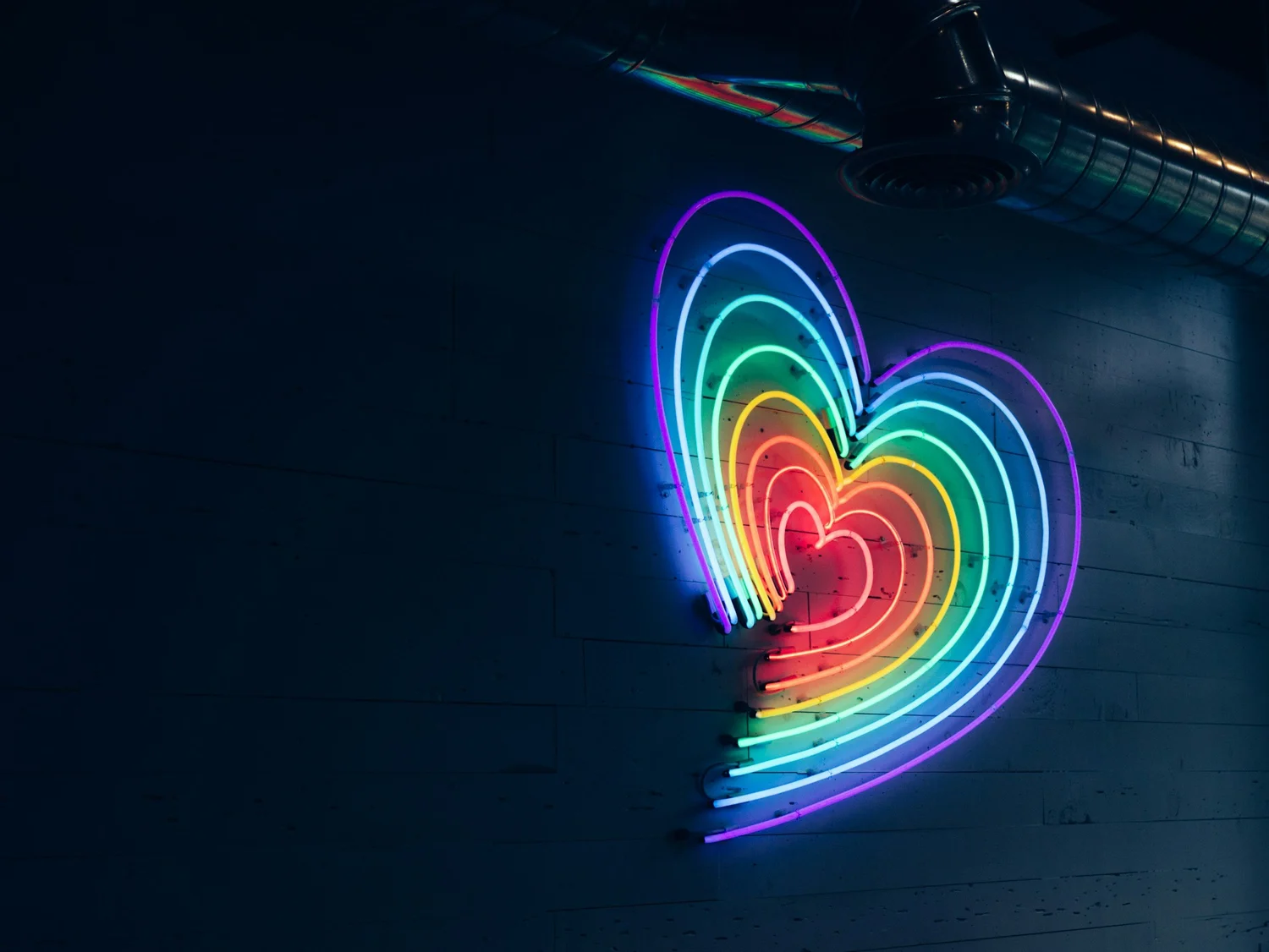 Rainbow-colored neon heart sign made of nested outlines glowing on a dark wall