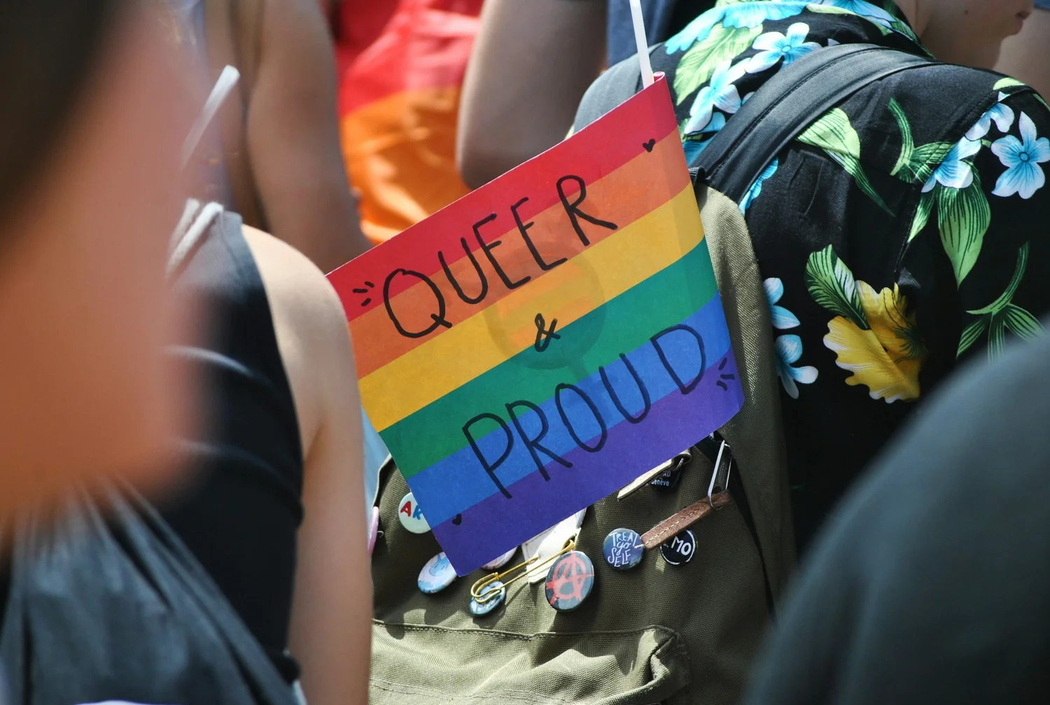 Handwritten rainbow sign reading "Queer & proud" in a crowd, in front of a backpack with pins