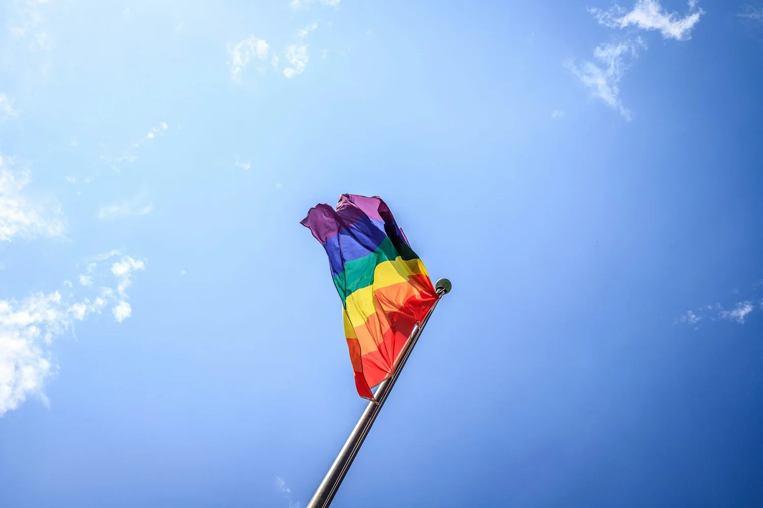 Rainbow flag waving on a flagpole against a blue sky with thin clouds