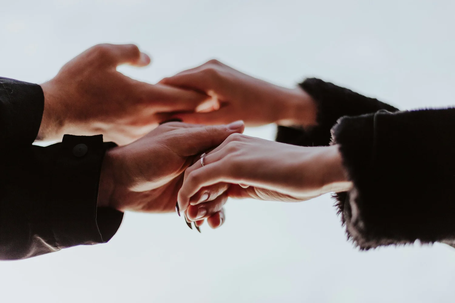 Four hands from two people clasping and touching fingers against a pale gray background