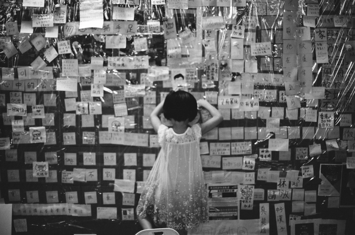 Black-and-white scene of a person in a dress seen from behind, hands on head, facing a wall covered in paper notes