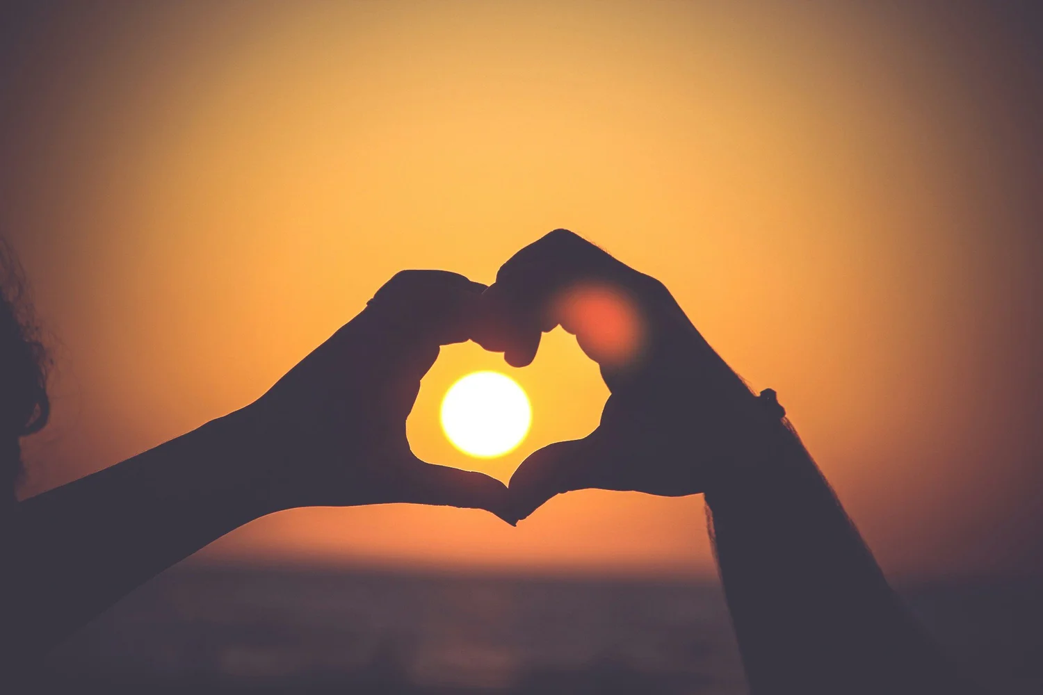 Silhouetted hands forming a heart shape around the sun in an orange sky