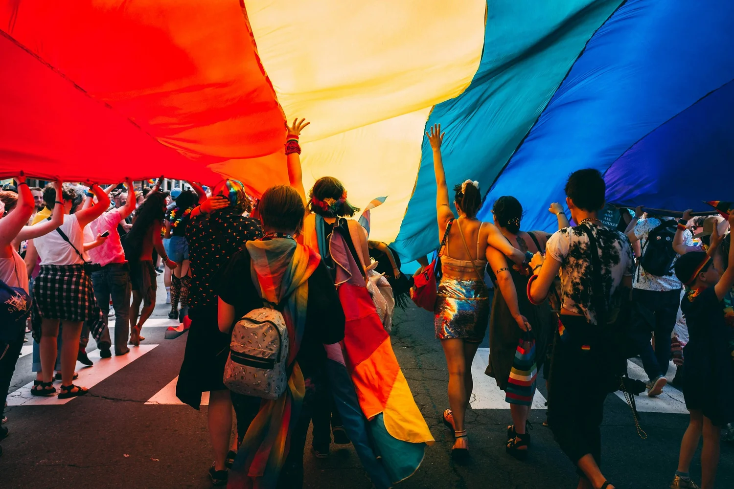 A crowd of people walking beneath a large rainbow flag held overhead, some wearing rainbow flags as capes