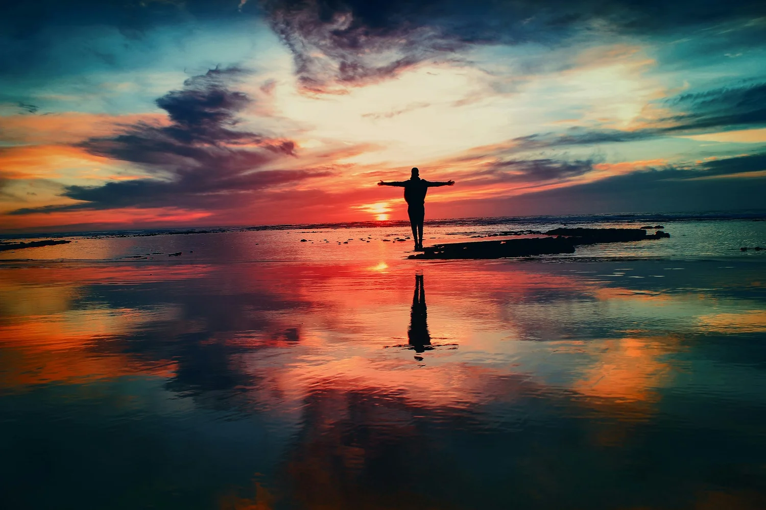 Silhouette of a person with arms outstretched on wet sand, the orange and blue sky reflected around them