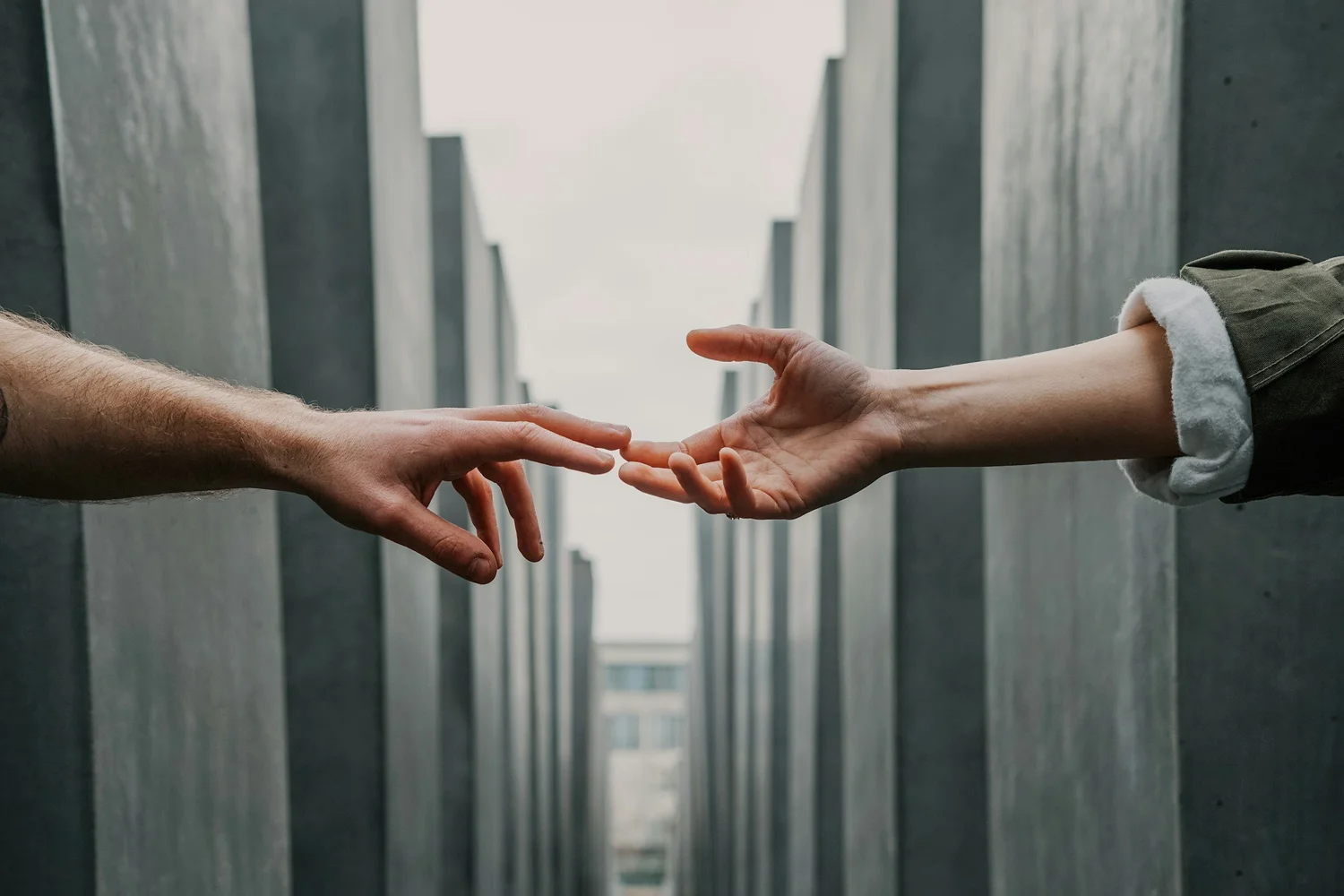 Two hands reaching toward each other, fingertips nearly touching, between tall gray concrete blocks