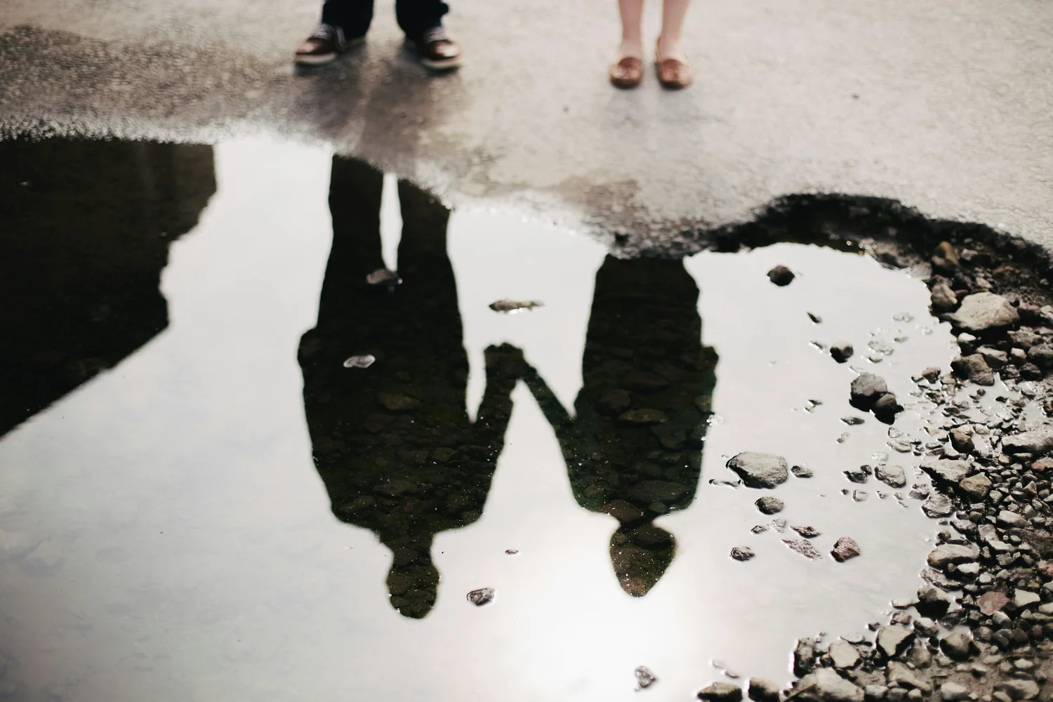 Two people holding hands, seen as a reflection in a puddle, with their feet at the top edge
